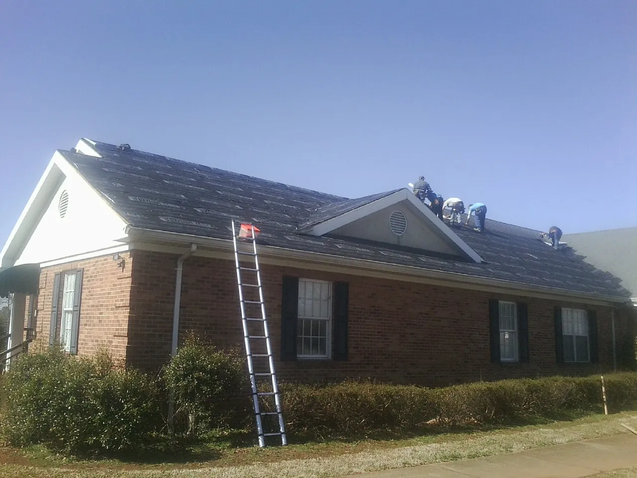 Crew tearing shingles off a brick ranch roof with a ladder set — Metal Roof Repair work by our crew in Tryon