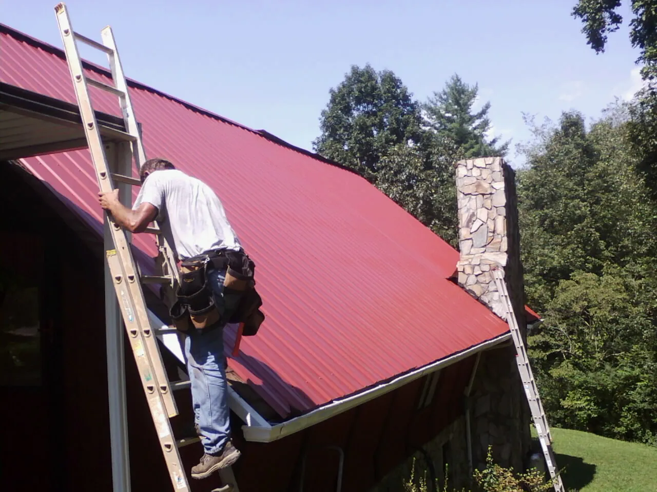 Roofer on a ladder beside a red metal roof and stone chimney — Soffit & Fascia Repair by Roofing Brothers in Tryon